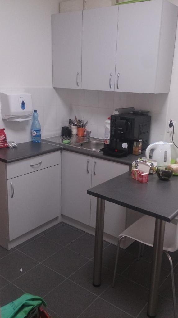 Niewielka kuchnia biurowa z szarymi szafkami i blatem, wyposażona w ekspres do kawy, czajnik elektryczny i akcesoria kuchenne, widoczny stół z krzesłem.