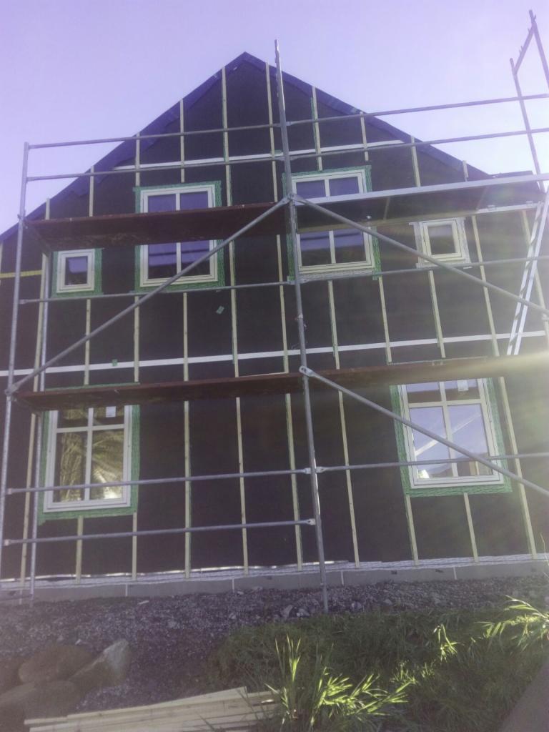 Wznoszenie ściany szkieletowej domu z widocznymi oknami, pokrytej czarną membraną i drewnianymi listwami, z rusztowaniem na pierwszym planie.