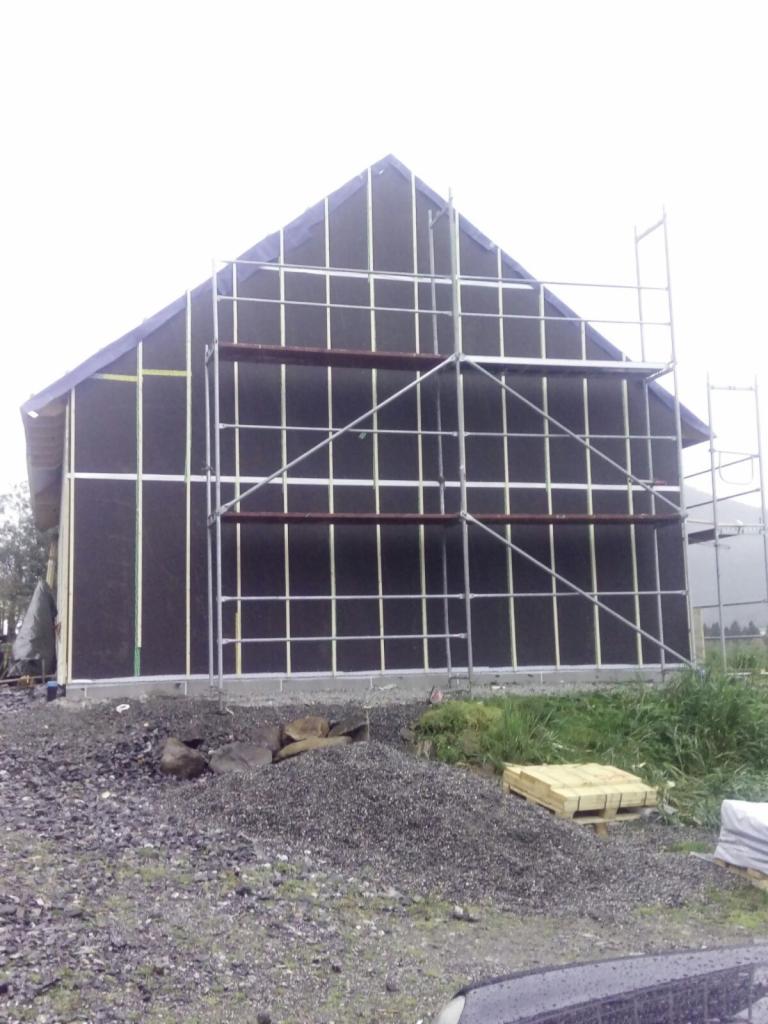 Budowa domu: widok na szkieletową konstrukcję z rusztowaniem, czarną membraną i drewnianymi belkami, na pierwszym planie gruz i paleta.