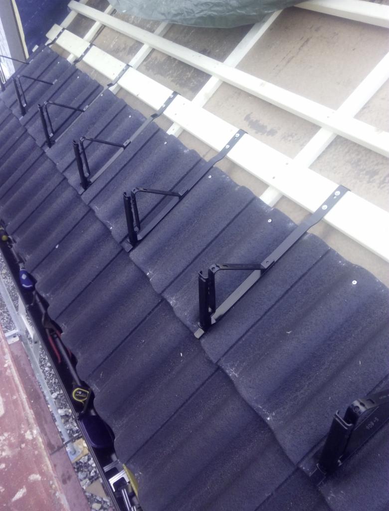 Montaż czarnych wsporników na szarym dachu z falistej blachy, widoczne narzędzia w rynnie i fragment drewnianej konstrukcji dachu.