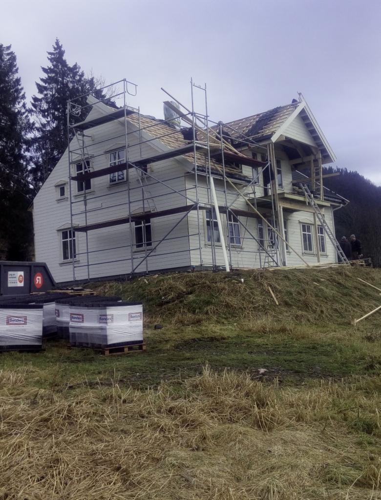 Dwu-kondygnacyjny dom szkieletowy w trakcie budowy, z widocznymi rusztowaniami i częściowo położonym dachem, materiały budowlane na paletach na pierwszym planie, pochmurne niebo w tle.