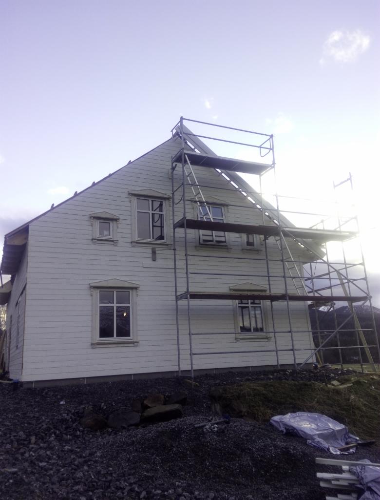 Wznoszony dom z białymi panelami i dekoracyjnymi obramowaniami okien, widoczne rusztowanie i teren przygotowany pod fundament.