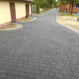 Szeroki, szary chodnik z drobnej kostki prowadzący między budynkami, z widoczną betoniarką w tle.