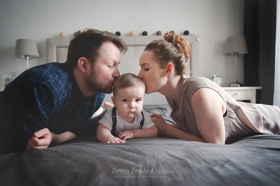 Rodzice całują w czoło niemowlę leżące na szarym łóżku; dziecko w białej koszulce i szelkach patrzy w obiektyw, w tle zagłówek z dekoracyjnymi kulkami.