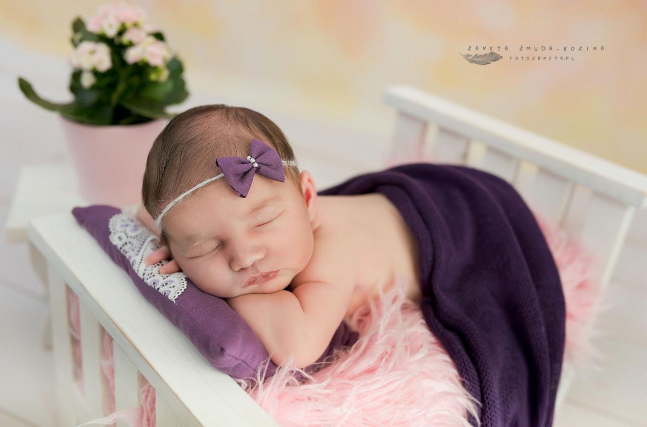 Śpiąca noworodek w fioletowym kokardką na głowie, leżąca na małym białym łóżeczku z fioletową poduszką i fioletowym kocykiem, obok różowa doniczka z kwiatami.