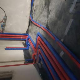 AQUAPIPE Dawid Bernat - Wiązka rur grzewczych w izolacji czerwono-niebieskiej, ułożona na czarnej folii izolacyjnej przy ścianie w trakcie montażu instalacji.
