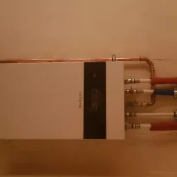 Biały, wiszący kocioł gazowy marki Buderus z widocznymi rurami miedzianymi i plastikowymi w kolorach czerwonym i niebieskim, zamontowany na beżowej ścianie.