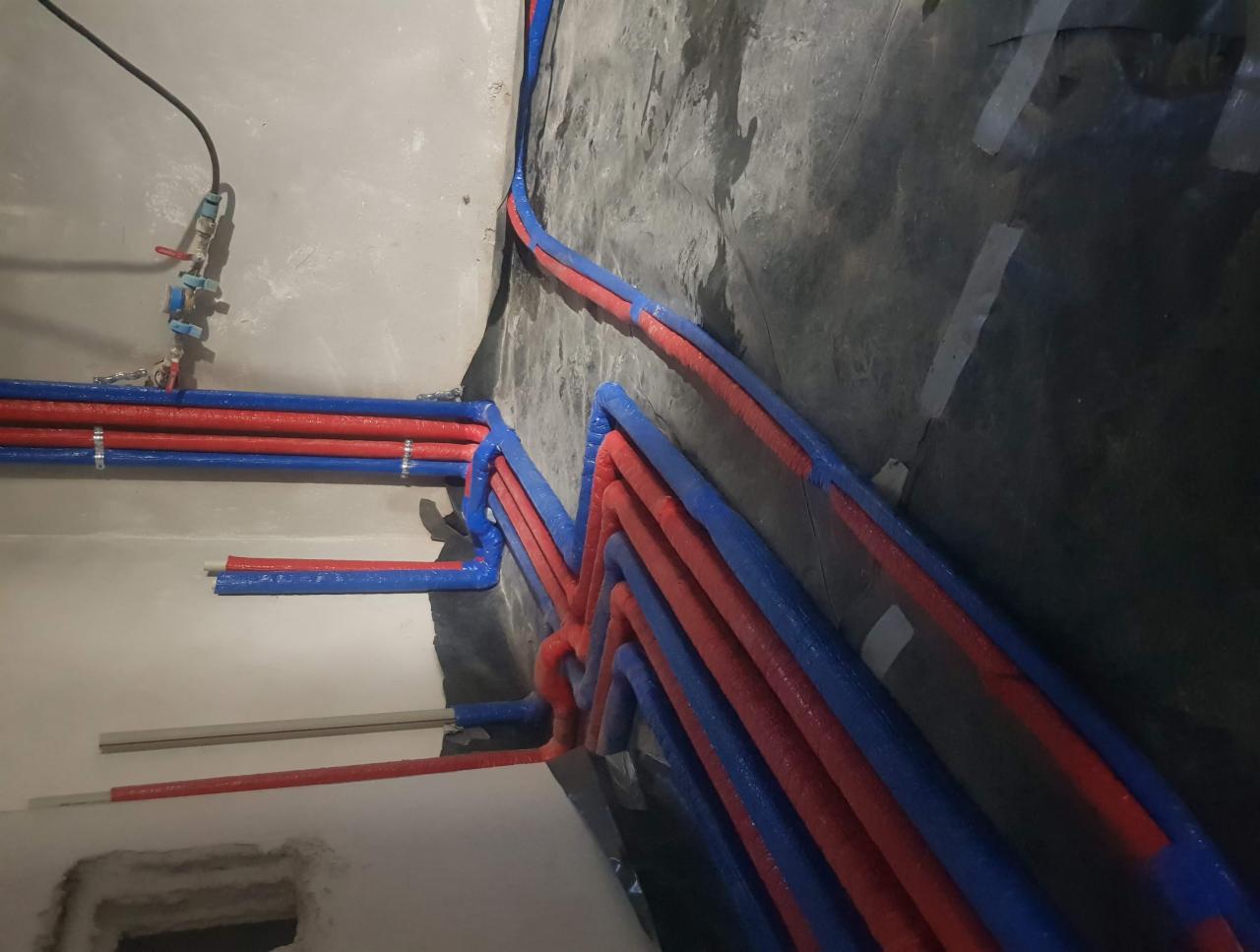 Wiązka rur grzewczych w izolacji czerwono-niebieskiej, ułożona na czarnej folii izolacyjnej przy ścianie w trakcie montażu instalacji.