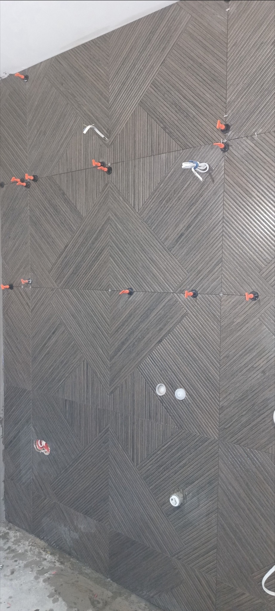 Ściana łazienki w trakcie wykańczania, pokryta brązowymi płytkami ułożonymi w geometryczny wzór, z widocznymi elementami poziomowania płytek, przewodami elektrycznymi i otworami na punkty świetlne.