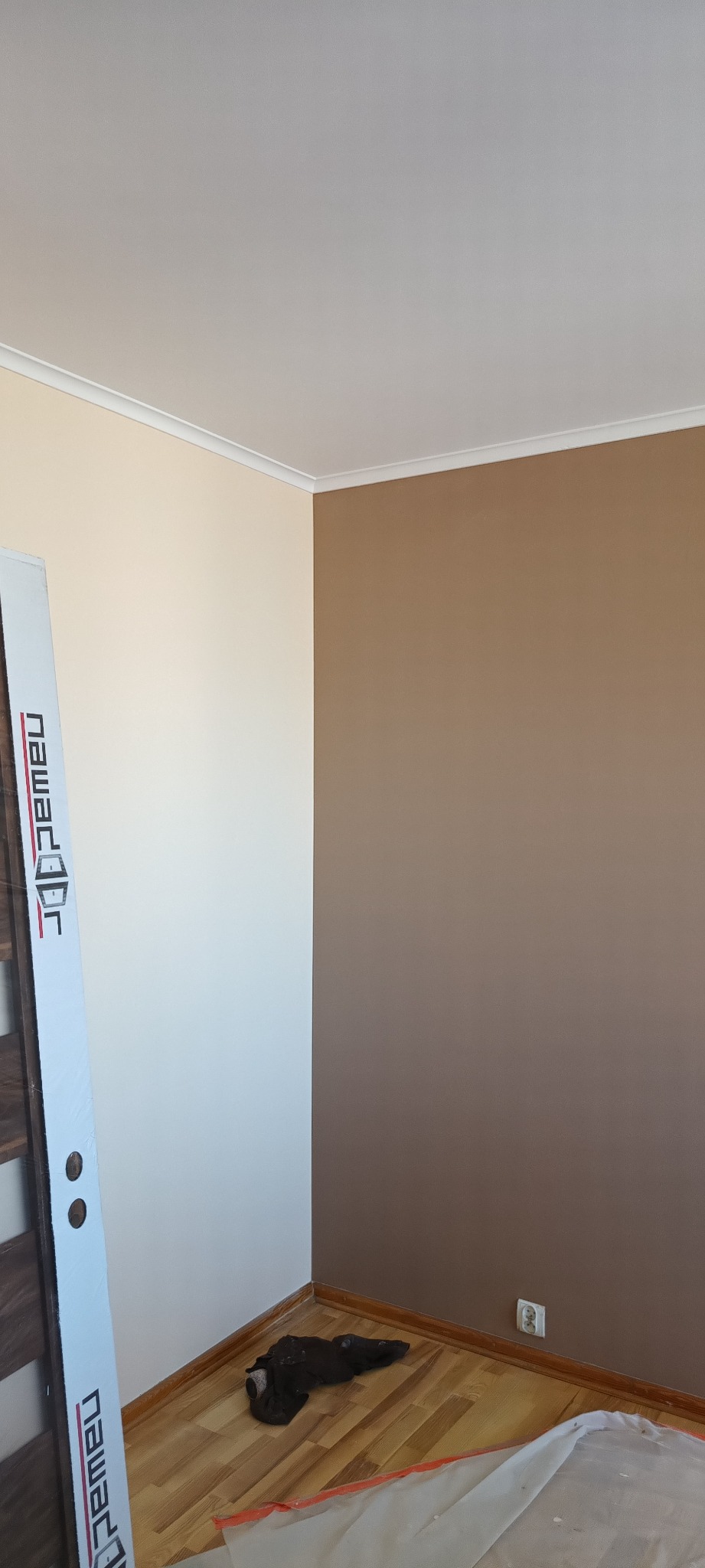 Pomieszczenie po malowaniu ścian na dwa kolory, z widocznym fragmentem drzwi w folii ochronnej, gniazdkiem elektrycznym i pozostałościami po pracy na podłodze.