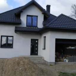 Dom jednorodzinny z jasną elewacją, czarnym dachem i stolarką okienną, widoczny stos piasku przed wejściem oraz otwarty garaż z materiałami budowlanymi w środku.