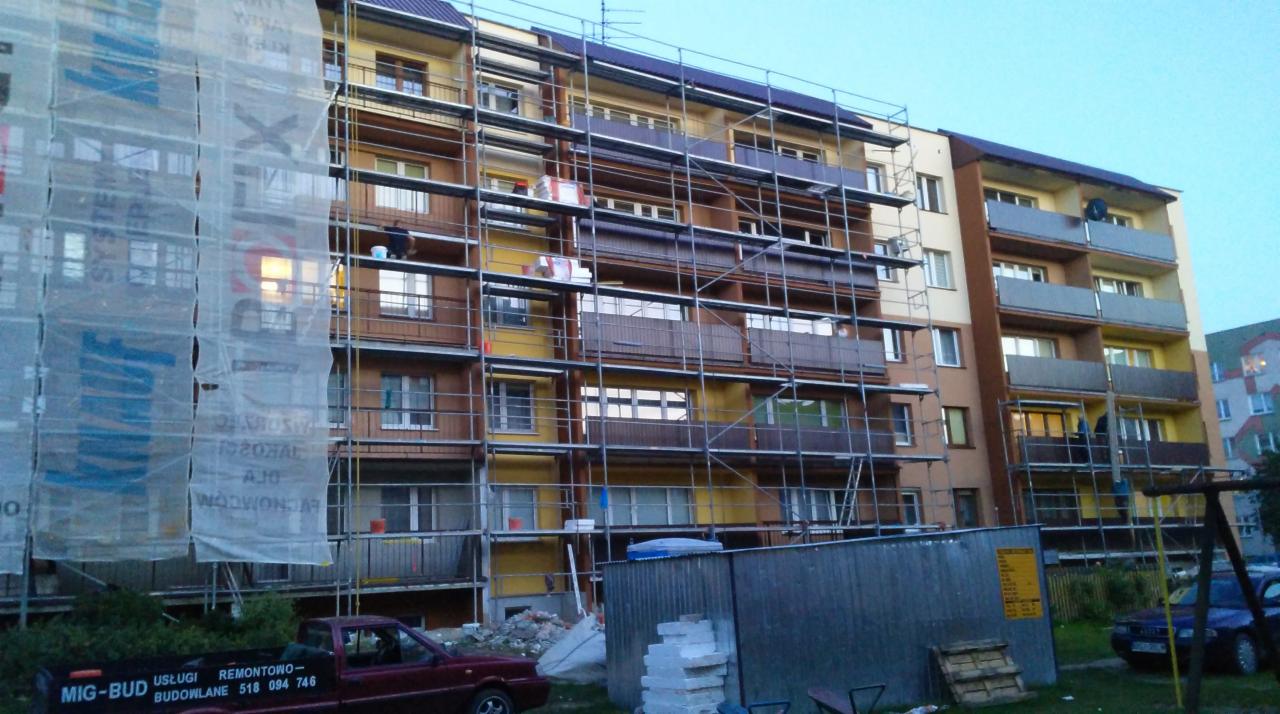 Wielopiętrowy budynek w trakcie renowacji elewacji, osłonięty siatką rusztowaniową z widocznymi rusztowaniami i pracownikiem przy oknie, na pierwszym planie zaparkowany samochód firmy budowlanej.