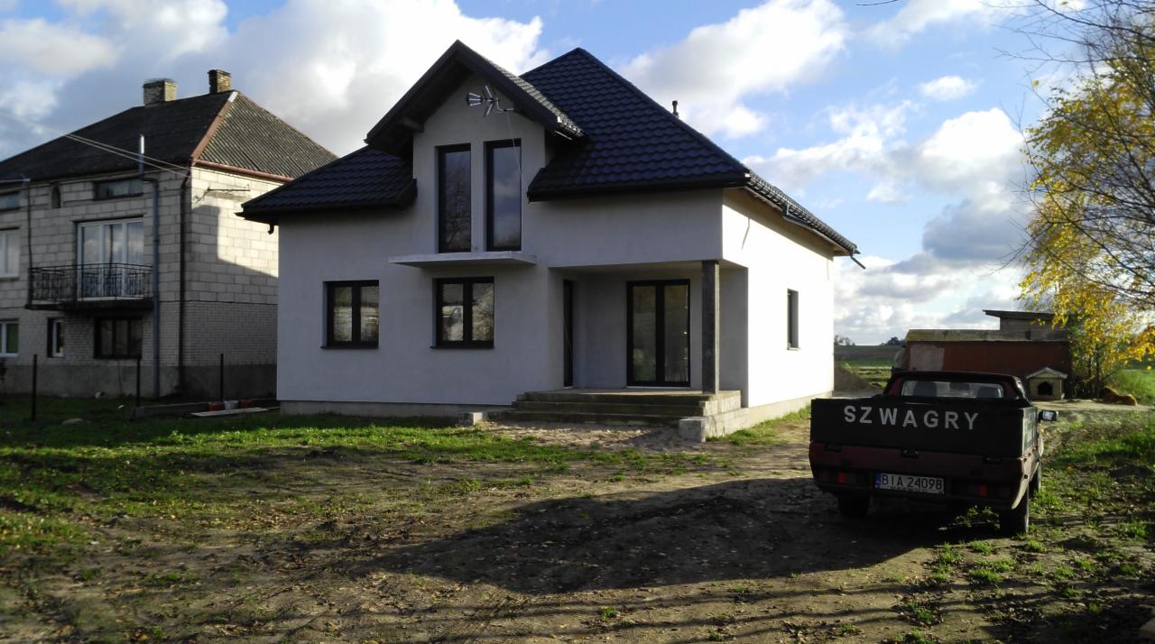 Nowoczesny dom jednorodzinny z szarą elewacją, ciemną stolarką okienną i czarnym dachem, widziany z zewnątrz na tle błękitnego nieba z chmurami, obok stoi czerwony pick-up z napisem 'SZWAGRY' na pace.