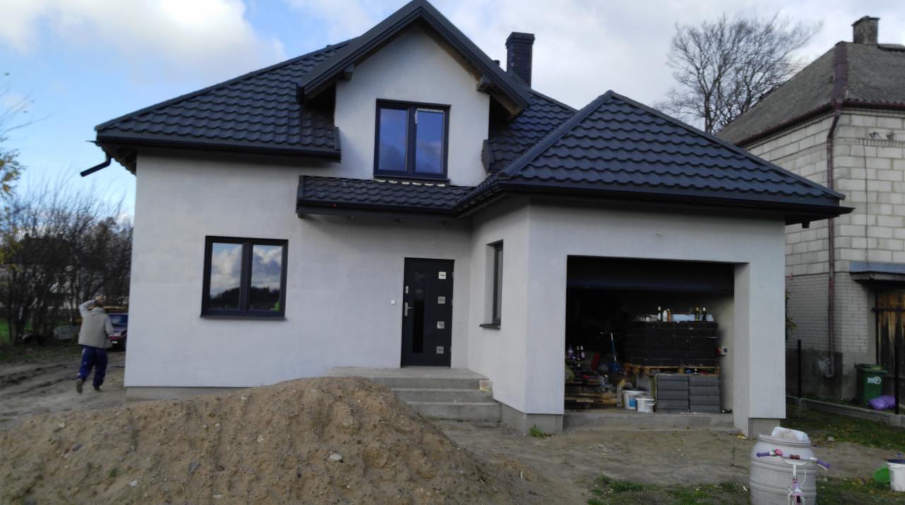 Dom jednorodzinny z jasną elewacją, czarnym dachem i stolarką okienną, widoczny stos piasku przed wejściem oraz otwarty garaż z materiałami budowlanymi w środku.