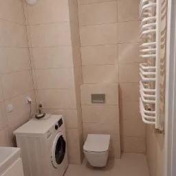 Beżowa łazienka z pralką Gorenje, wiszącą toaletą i grzejnikiem drabinkowym, wykończona płytkami w jednolitym kolorze.