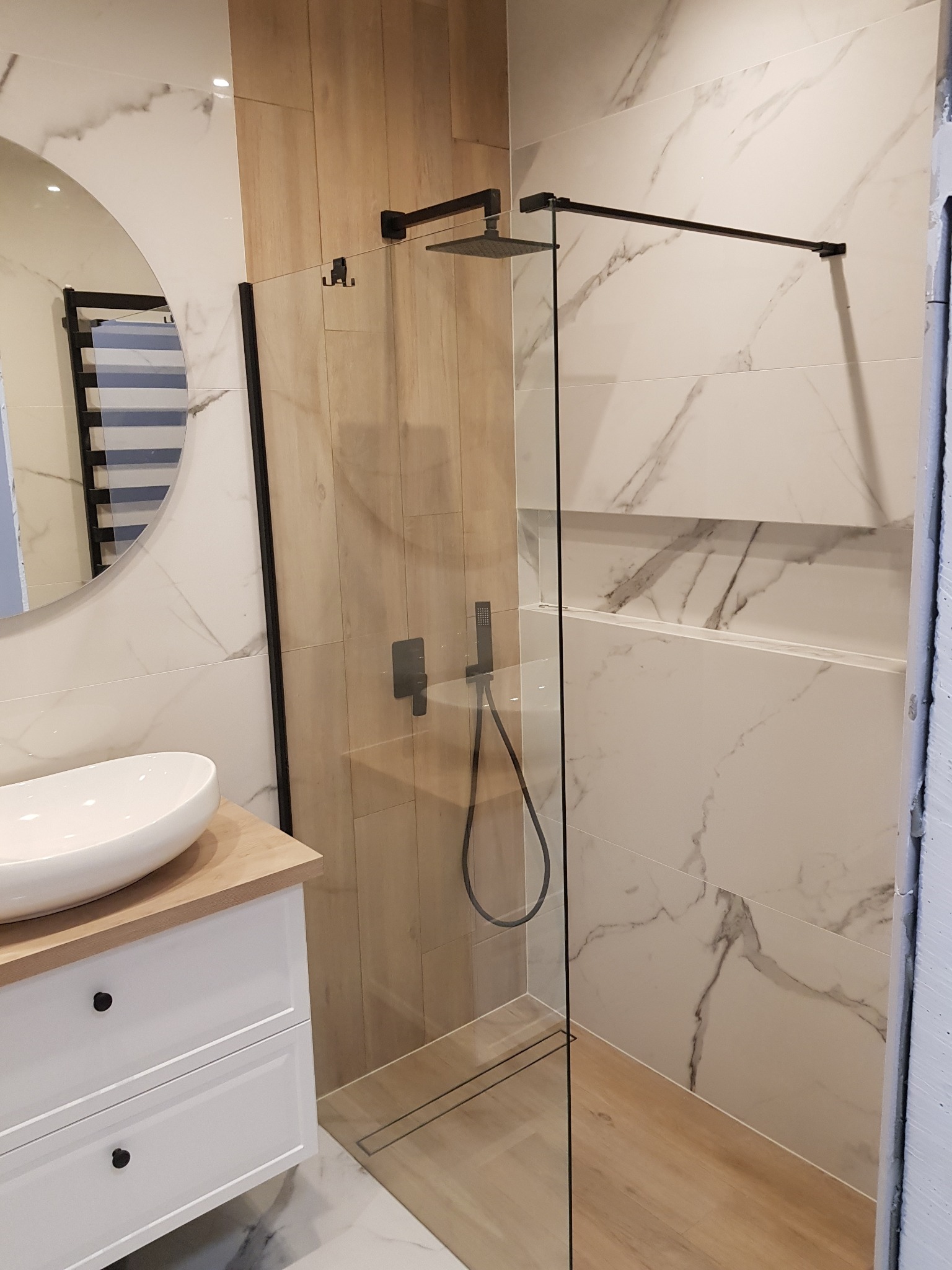Nowoczesna łazienka z kabiną prysznicową typu walk-in z czarną armaturą, umywalką nablatową na białej szafce z drewnianym blatem i grzejnikiem drabinkowym w okrągłym lustrze.
