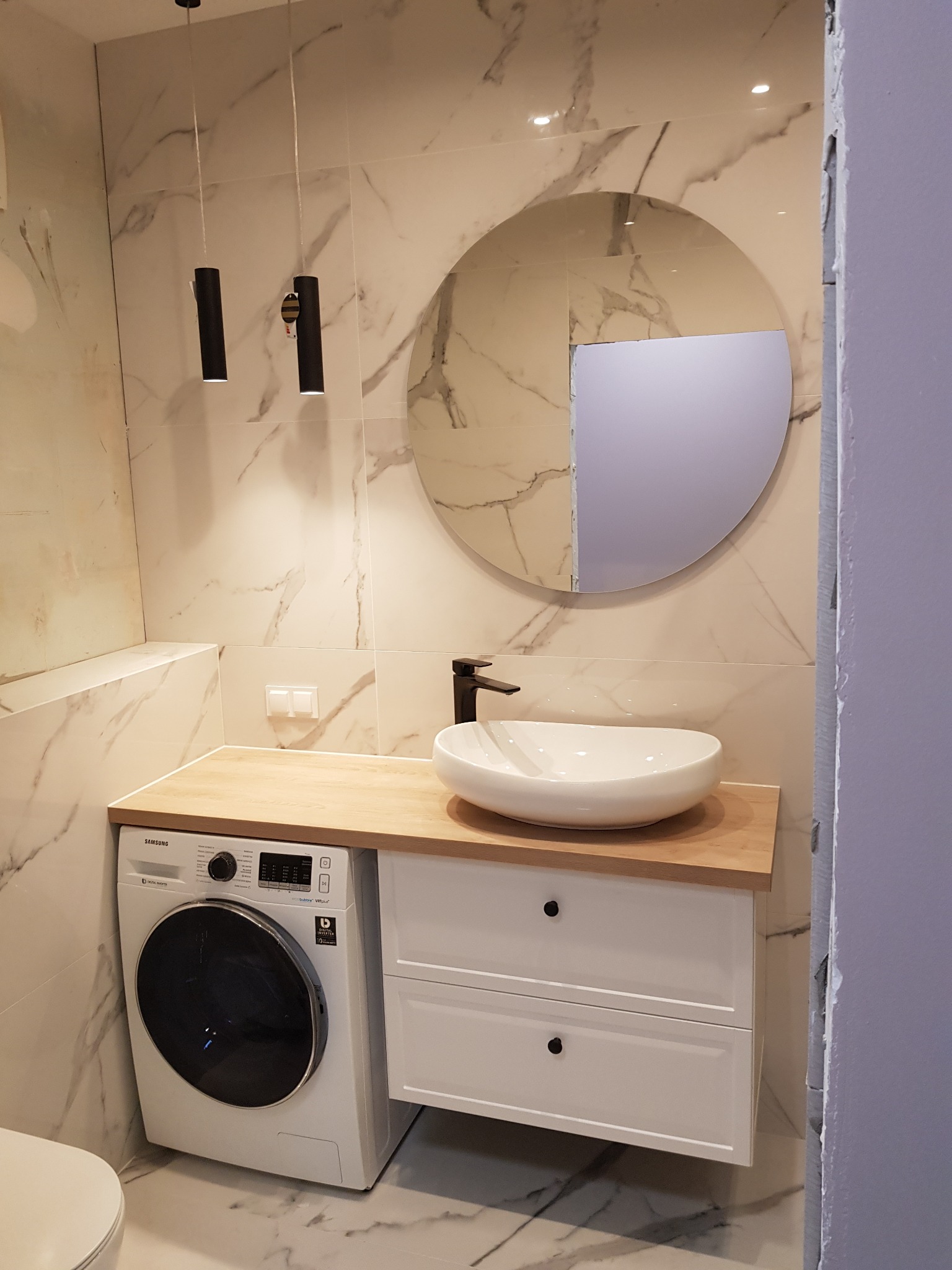 Nowoczesna łazienka z marmurowymi płytkami, okrągłym lustrem, białą umywalką nablatową na drewnianym blacie, podwieszaną szafką z dwiema szufladami i pralką automatyczną.
