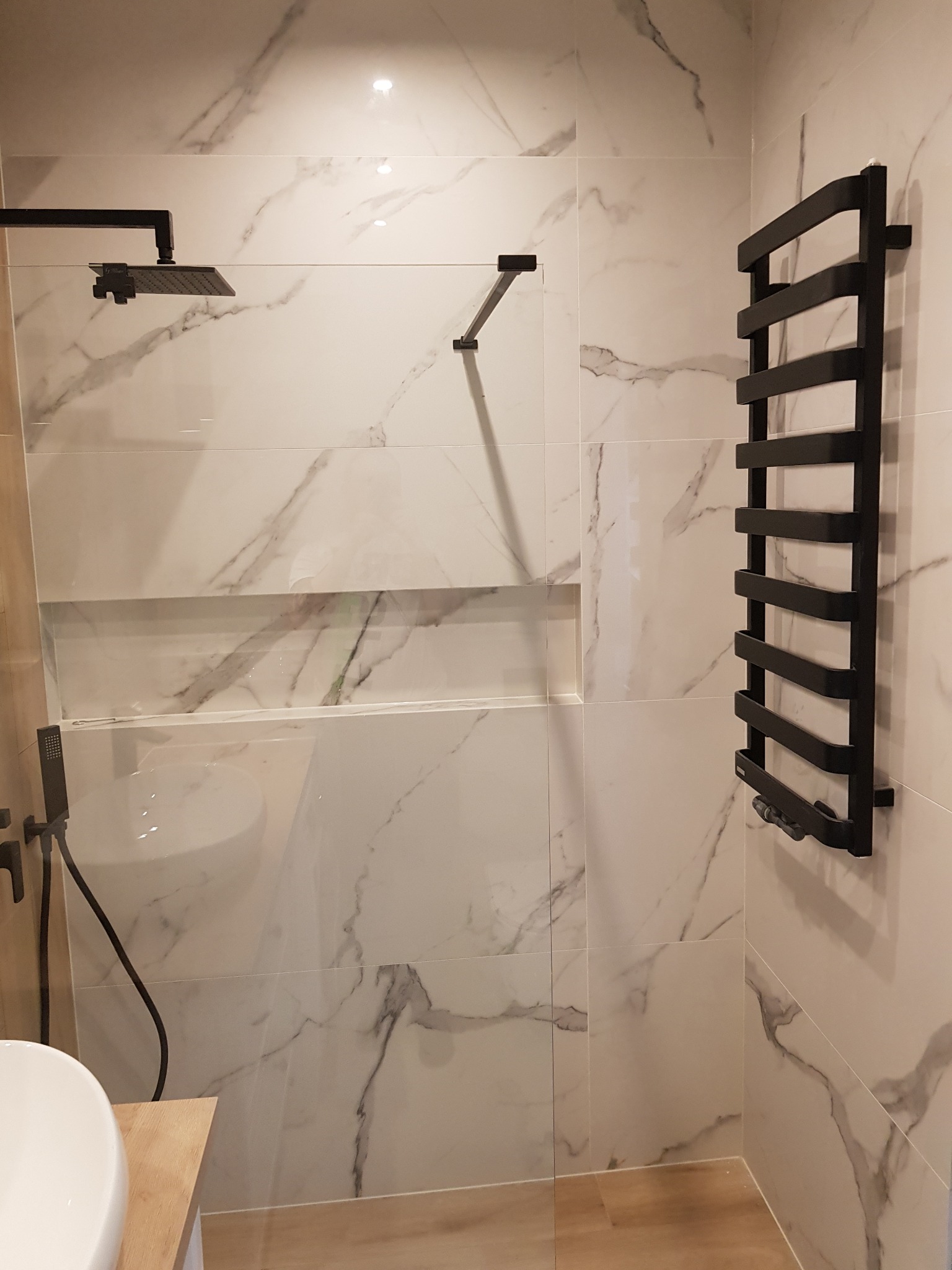 Nowoczesna łazienka z białymi płytkami imitującymi marmur, czarną armaturą i grzejnikiem drabinkowym, widoczny fragment umywalki i półka w ścianie.
