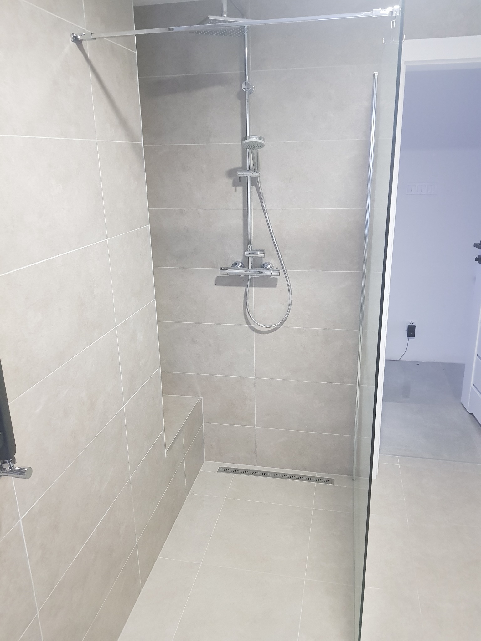 Nowoczesna kabina prysznicowa walk-in z odpływem liniowym w podłodze, wykończona płytkami w odcieniach szarości, z zamontowaną deszczownicą i słuchawką prysznicową.