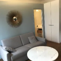 Jasnoszara sofa z poduszkami w salonie z okrągłym stolikiem marmurowym, na ścianie dekoracyjne lustro w kształcie słońca, w tle biała szafa wnękowa i otwarte drzwi do łazienki.