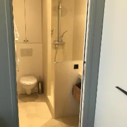 Widok na minimalistyczną łazienkę z kabiną prysznicową typu walk-in, toaletą podwieszaną i szafkami w zabudowie, widzianą zza uchylonych drzwi szafy w kolorze złamanej bieli.