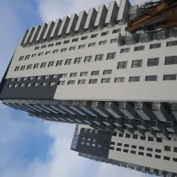 Wielopiętrowy budynek z białymi i szarymi panelami elewacyjnymi, widok z dołu na tle błękitnego nieba z chmurami, ramię dźwigu widoczne w prawym górnym rogu.