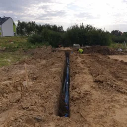 Wykop z ułożonymi niebieskimi rurami, widoczny pracownik w odblaskowej kamizelce i geodezyjny niwelator w tle, teren budowy.