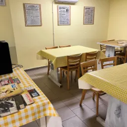 Jasne wnętrze kawiarni z drewnianymi krzesłami i stolikami nakrytymi obrusami w żółtą kratę. Na ścianach wiszą oprawione gazety. Na stole leżą gazety i stoi ekspres do kawy.