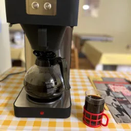 Ekspres Hendi z kawą na stole w żółtą kratę, obok szklanka z uchwytem i gazeta. Widok w jadalni, prawdopodobnie w trakcie przygotowań do cateringu.