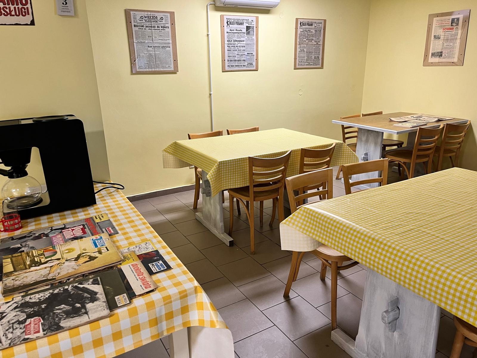 Jasne wnętrze kawiarni z drewnianymi krzesłami i stolikami nakrytymi obrusami w żółtą kratę. Na ścianach wiszą oprawione gazety. Na stole leżą gazety i stoi ekspres do kawy.