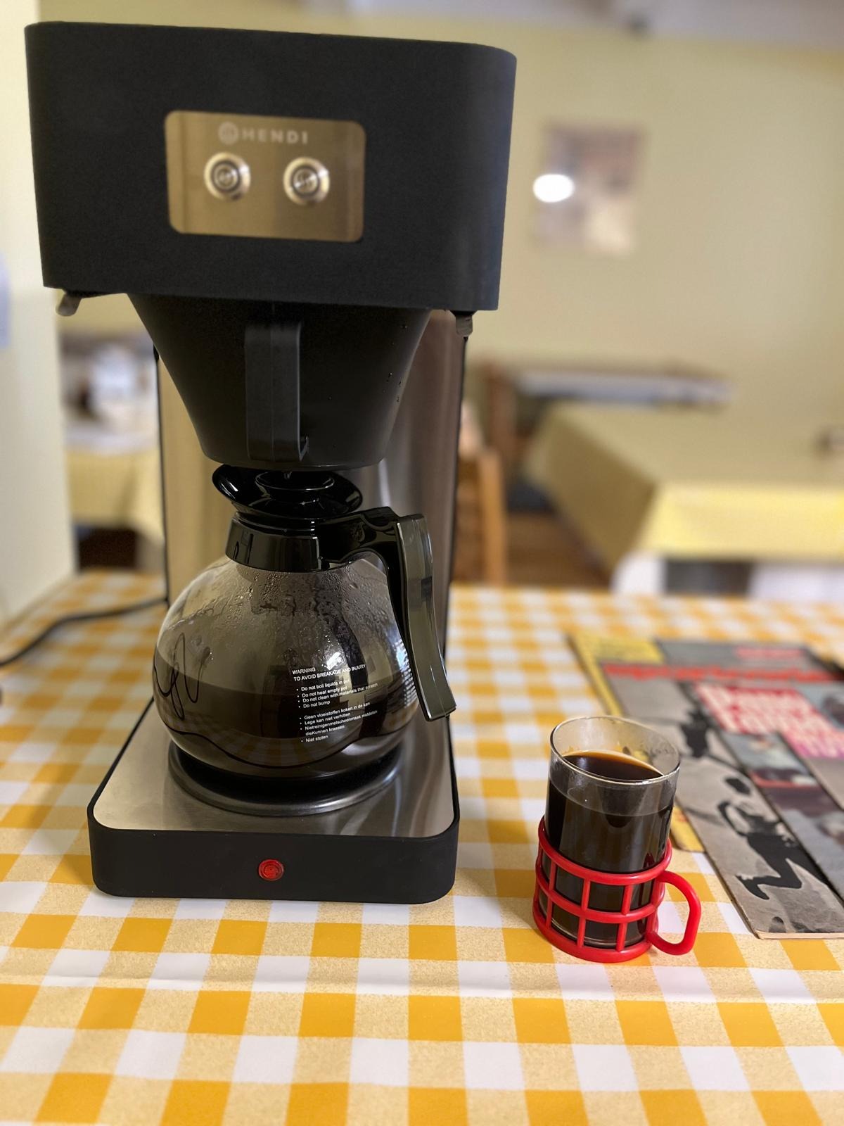 Ekspres Hendi z kawą na stole w żółtą kratę, obok szklanka z uchwytem i gazeta. Widok w jadalni, prawdopodobnie w trakcie przygotowań do cateringu.