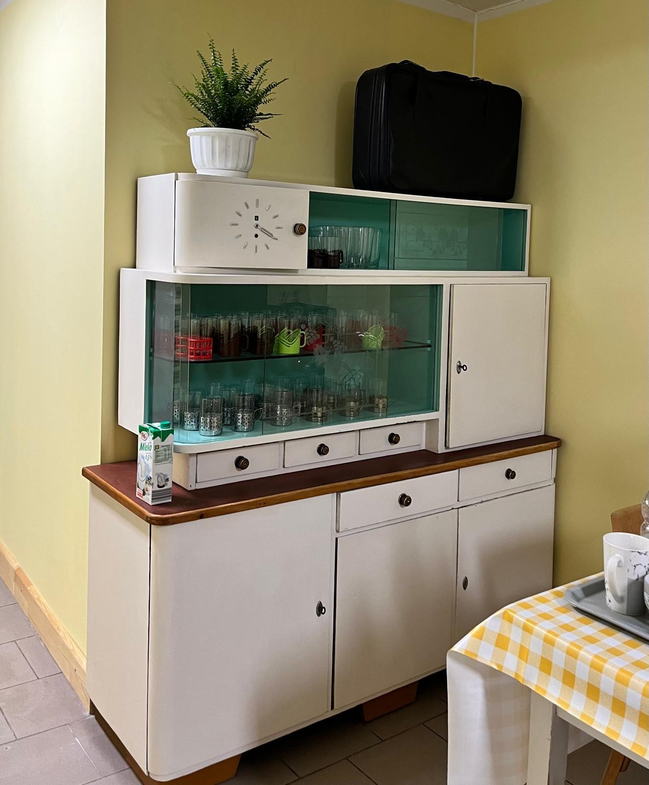 Retro kredens w salonie z rośliną i walizką na górze. W szklanych witrynach widoczne szklanki i filiżanki. Na blacie karton mleka. Żółte ściany w tle.