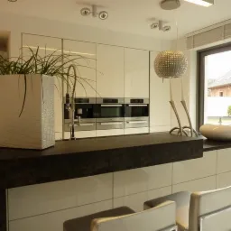 Nowoczesna kuchnia z białymi, lakierowanymi frontami, ciemnym blatem i wyspą kuchenną z hokerami. Na wyspie stoi duża, biała donica o fakturze skóry krokodyla. Widoczne wbudowane sprzęty AGD...