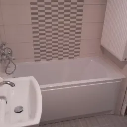 Wykończona łazienka z białą wanną, umywalką z chromowaną baterią i grzejnikiem na ścianie obłożonej płytkami w szaro-beżową szachownicę.