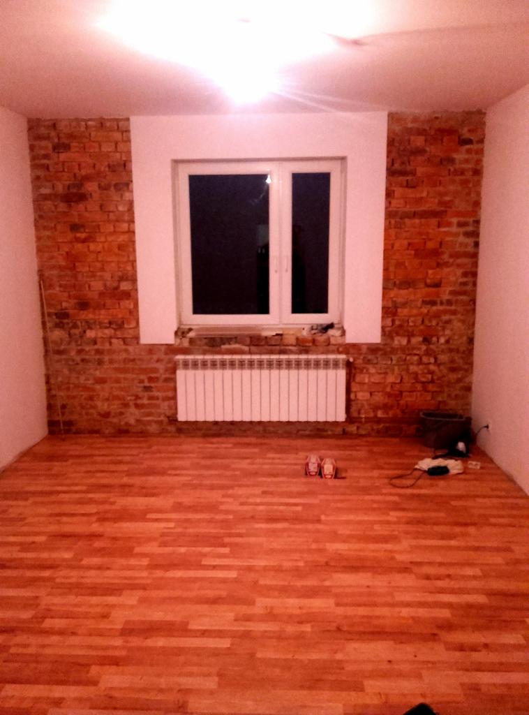 Wnętrze pokoju w Żmigrodzie po remoncie, z nową drewnianą podłogą, fragmentem ściany z cegły wokół okna i białym grzejnikiem pod nim. Widoczne ślady po remoncie i narzędzia.
