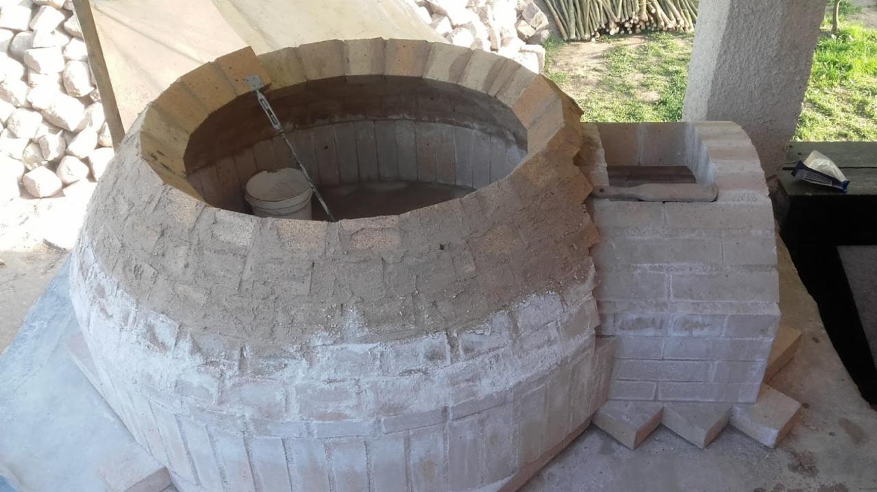 Budowa pieca tandoor z cegły, widoczny etap murowania i wykończenia zewnętrznego, z wiaderkiem i narzędziami wewnątrz, na zewnątrz widoczny stos gałęzi.
