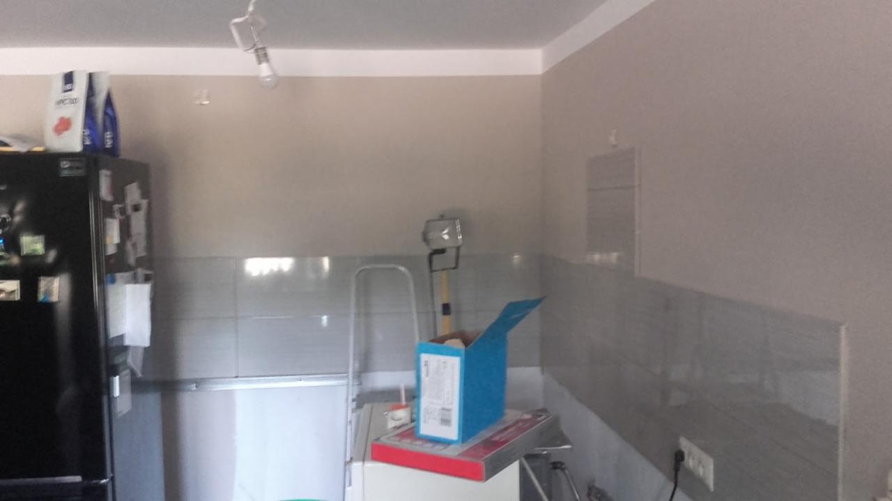 Świeżo pomalowane ściany w kuchni w odcieniach szarości, z widocznymi płytkami nad blatem, drabina i lampa halogenowa ustawione do dalszych prac wykończeniowych, kartonowe pudło i opakowania...