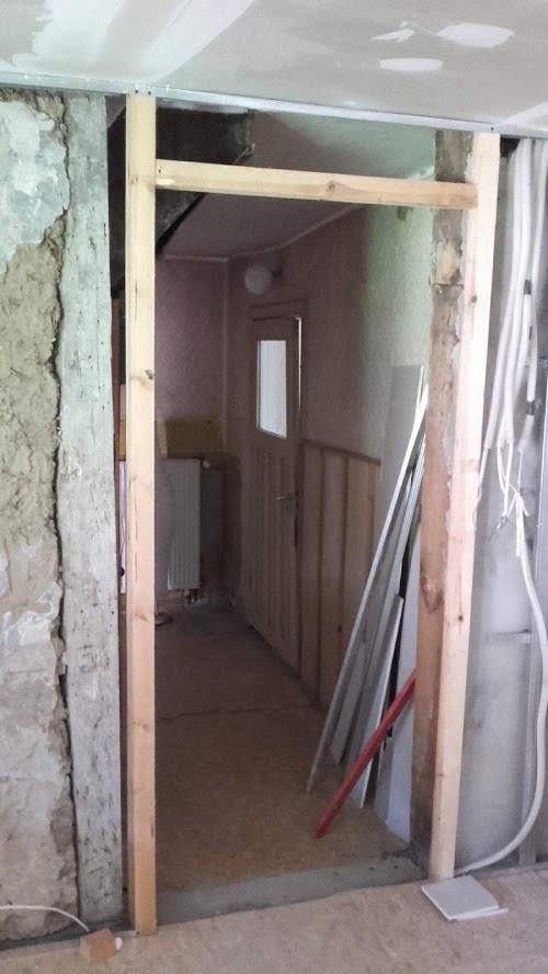 Wnętrze w trakcie prac remontowych: surowa ściana z odsłoniętą strukturą, drewniana rama drzwiowa w trakcie montażu, widoczne instalacje elektryczne i rury, wstępny etap prac wykończeniowych...