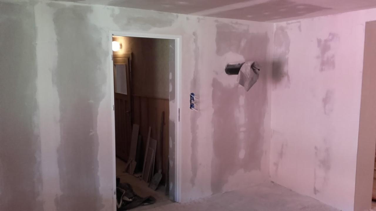 Pokój w trakcie szpachlowania ścian, widoczne niedoskonałości i pozostałości po gładzi, rury wystające ze ściany zabezpieczone szmatką, w tle otwarte drzwi do pomieszczenia z boazerią.