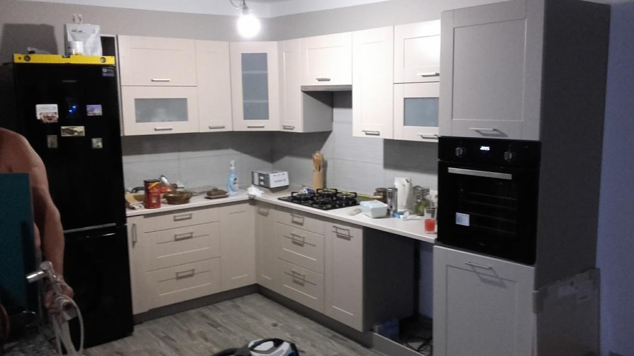 Jasna kuchnia z frontami w odcieniach beżu i szarości, czarną lodówką z żółtą poziomicą na górze i czarnym piekarnikiem w zabudowie, widok na część pomieszczenia podczas montażu.