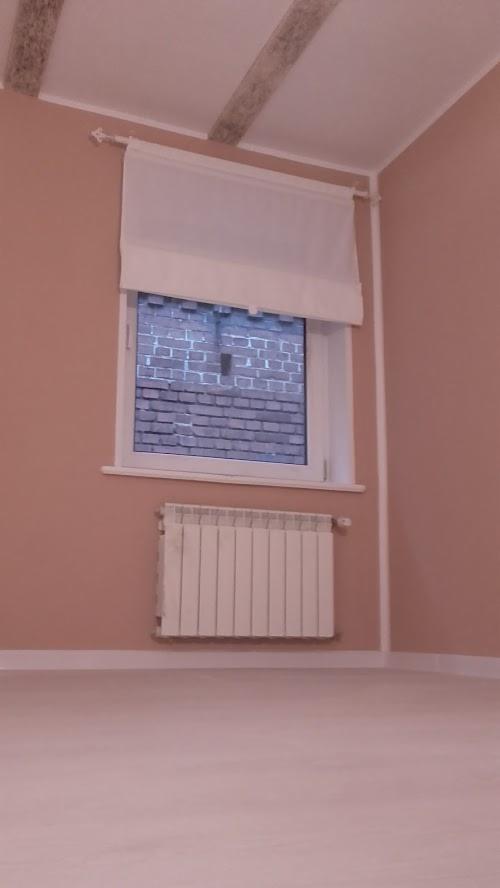 Wnętrze pokoju po remoncie: ściany w odcieniach różu, białe listwy przypodłogowe, grzejnik pod oknem z roletą rzymską, drewniane belki na suficie.