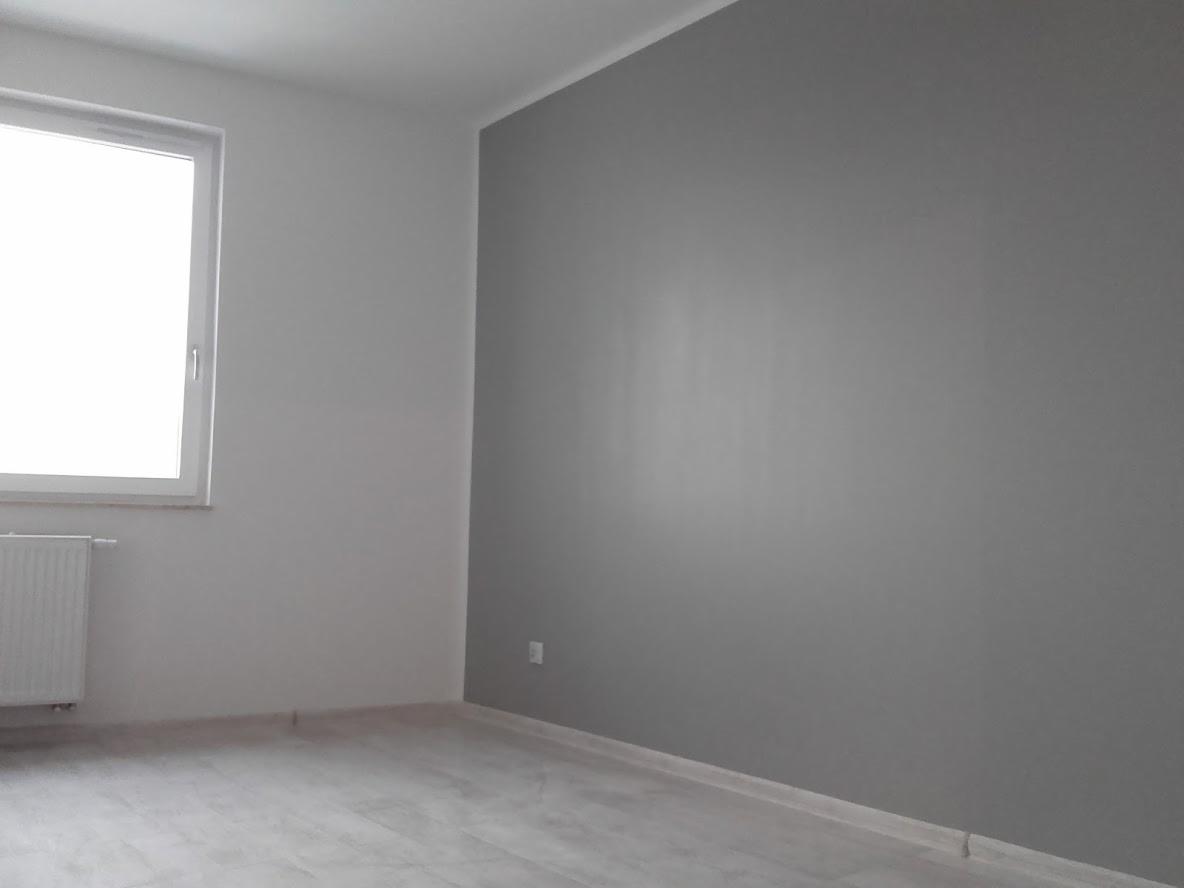 Pomalowane na szaro wnętrze pokoju z białym oknem i grzejnikiem, widoczne panele podłogowe imitujące drewno.