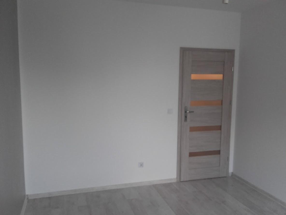 Pusty pokój po remoncie, ściany pomalowane na biało, szare panele podłogowe, jasnoszare drzwi z przeszkleniami i srebrną klamką.