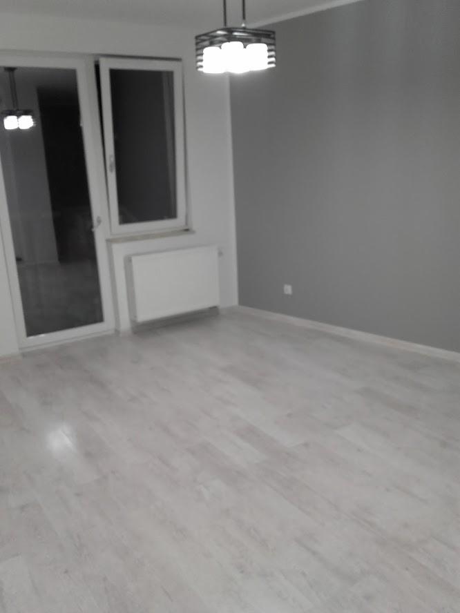 Pusty pokój po remoncie z jasną podłogą imitującą drewno, szarą ścianą, białym grzejnikiem i nowoczesną lampą sufitową z żarówkami LED.