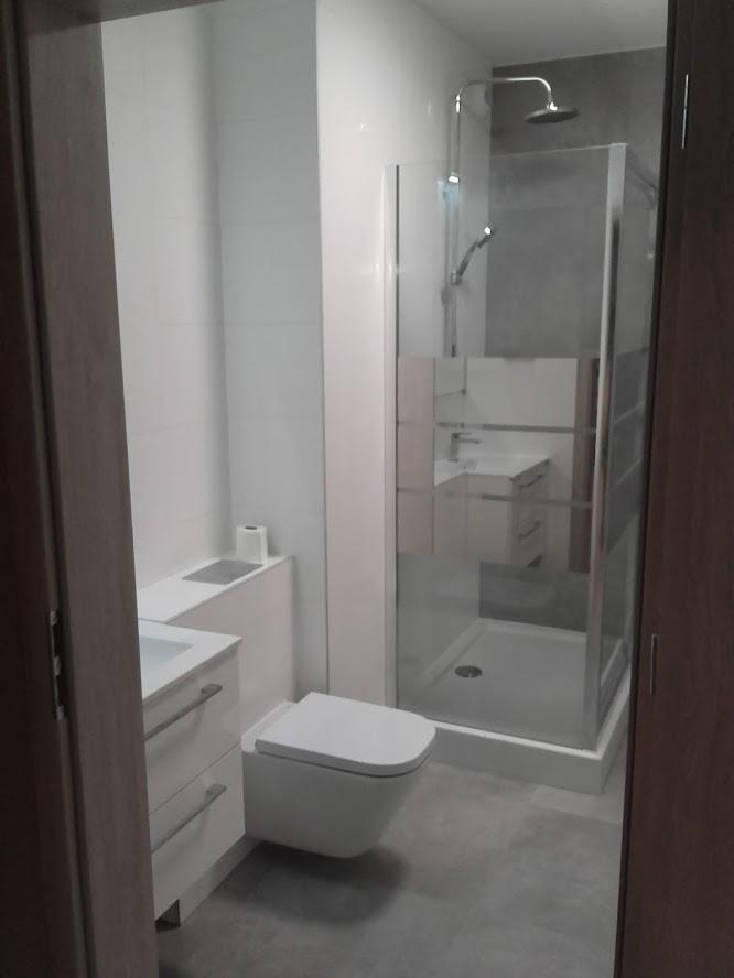 Nowoczesna łazienka po remoncie: biała ceramika, szara podłoga, kabina prysznicowa z deszczownicą, minimalistyczna szafka pod umywalką i toaleta podwieszana z ukrytą spłuczką.