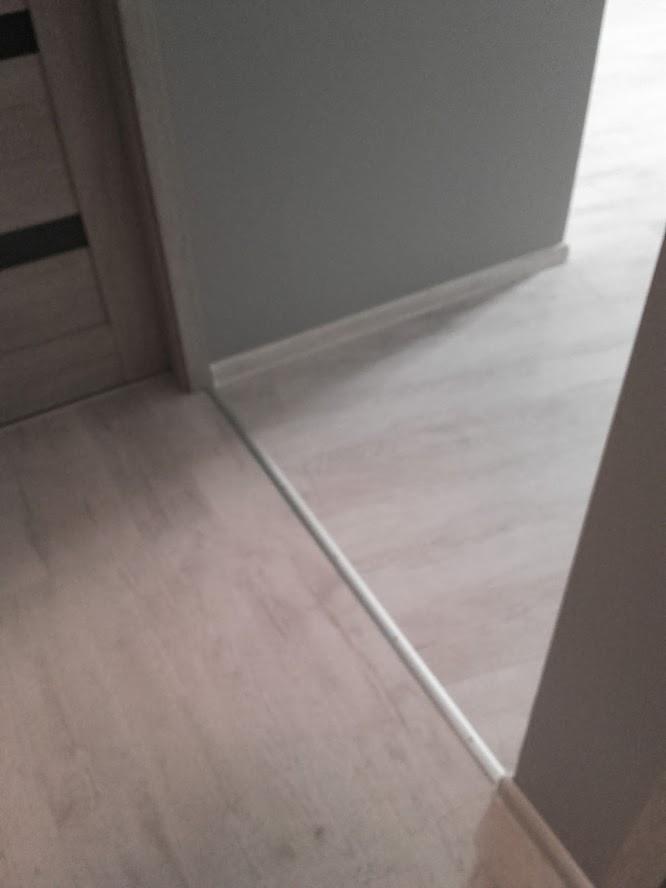 Szara ściana z białą listwą przypodłogową i panele podłogowe w odcieniach szarości, widok z bliska na łączenie ściany i podłogi przy drzwiach.