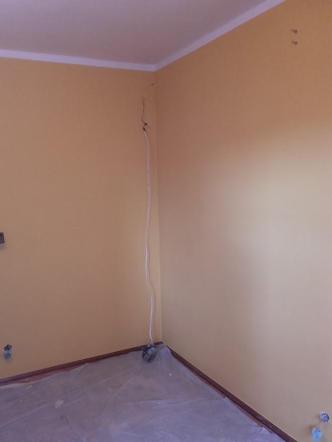 Pokój w trakcie malowania na żółto, z zabezpieczoną folią podłogą i widocznym przewodem elektrycznym wystającym ze ściany.