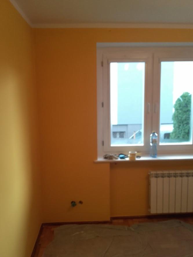 Pokój po malowaniu ścian na kolor musztardowy, z widocznym zakrytym podłogą folią malarską, kaloryferem i oknem z widokiem na zieleń.