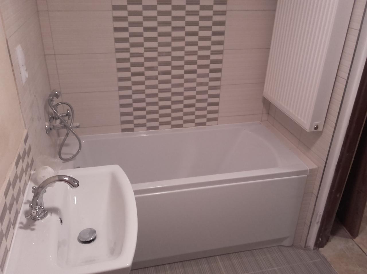 Wykończona łazienka z białą wanną, umywalką z chromowaną baterią i grzejnikiem na ścianie obłożonej płytkami w szaro-beżową szachownicę.