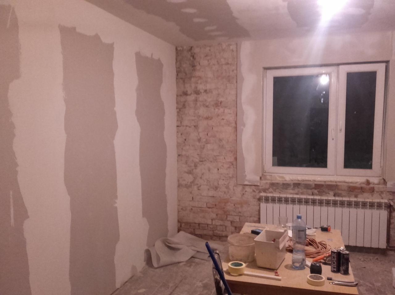 Wnętrze pokoju w trakcie remontu: ściany pokryte częściowo gładzią, odsłonięta cegła przy oknie, narzędzia i materiały na stole w centrum.